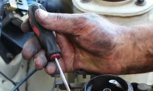Brudna ręka mechanika trzyma śrubokręt, naprawiając linkę zmiany biegów. Sprawdź wymiana linki zmiany biegów koszt.