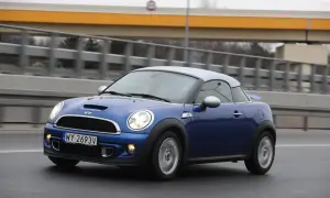 Niebieski Mini R58 Cooper S z szarym dachem pędzi po drodze, za nim widać most.
