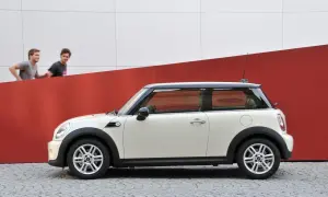 Biały Mini Cooper r56 z czarnym dachem stoi na bruku. Dwóch mężczyzn biegnie za nim na tle czerwono-białej ściany.