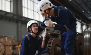 Dwóch pracowników w kombinezonach i kaskach dyskutuje przy użyciu krótkofalówki. To pokazuje, kim jest logistyk – osoba dbająca o sprawny przepływ towarów.