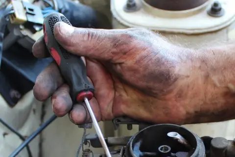 Brudna ręka mechanika trzyma śrubokręt, naprawiając linkę zmiany biegów. Sprawdź wymiana linki zmiany biegów koszt.