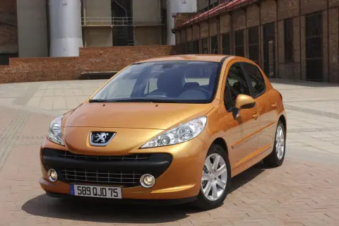 Pomarańczowy Peugeot 207 SW 1.4 benzyna zaparkowany na brukowanym placu przed ceglanym budynkiem.