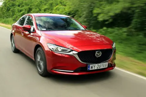 Czerwona Mazda 6 2.0 benzyna pędzi po wiejskiej drodze, otoczona zielenią drzew.