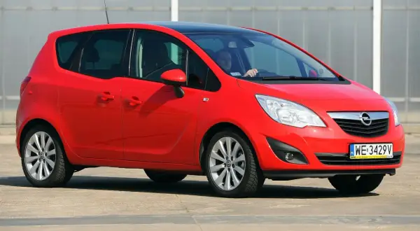 Czerwony Opel Meriva 1.6 benzyna z czarnym dachem, zaparkowany na zewnątrz. Kierowca w okularach siedzi za kierownicą.