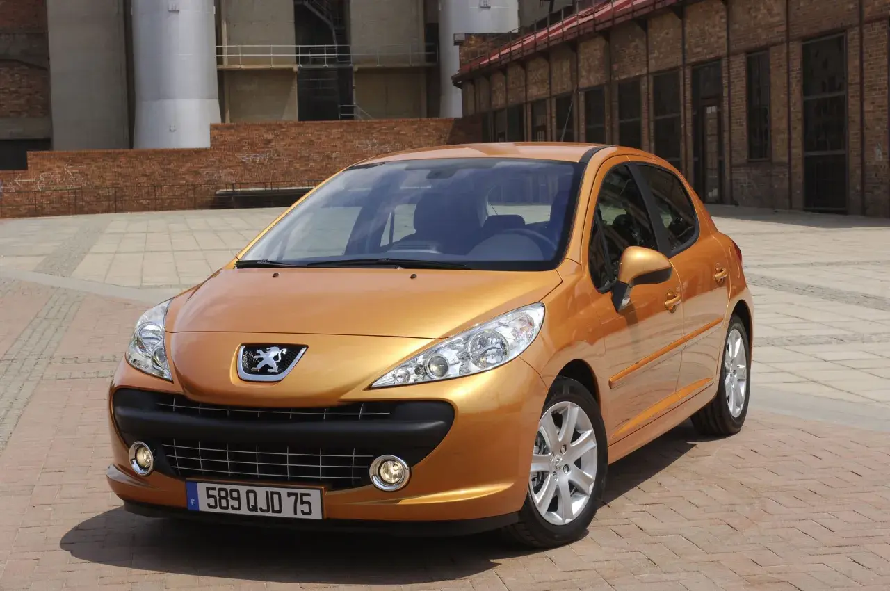 Pomarańczowy Peugeot 207 SW 1.4 benzyna zaparkowany na brukowanym placu przed ceglanym budynkiem.