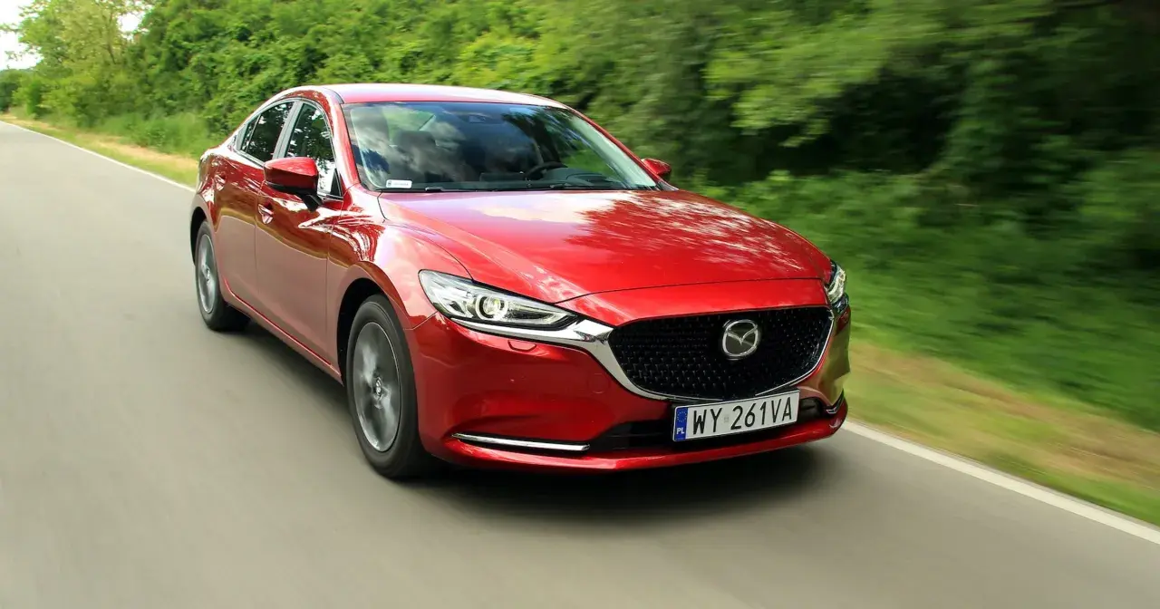 Czerwona Mazda 6 2.0 benzyna pędzi po wiejskiej drodze, otoczona zielenią drzew.