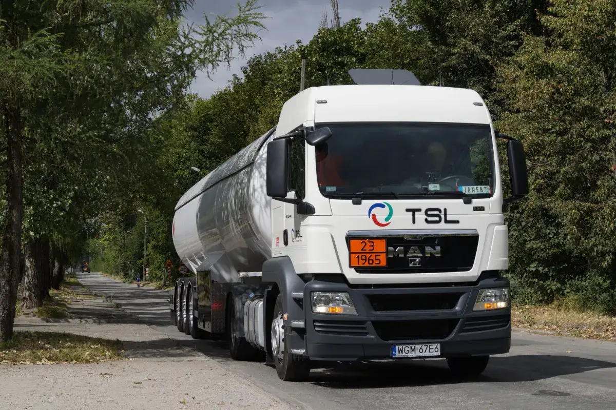 Biały cysterna z logo TSL jedzie drogą wśród drzew. To ważny element branży TSL, transportującej towary.