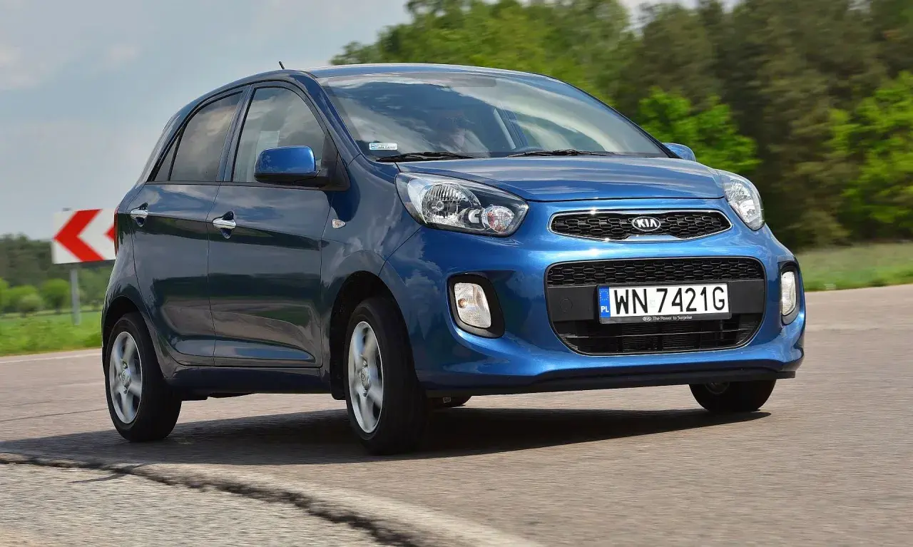 Niebieski Kia Picanto 2012 na drodze, z zielonymi drzewami w tle.