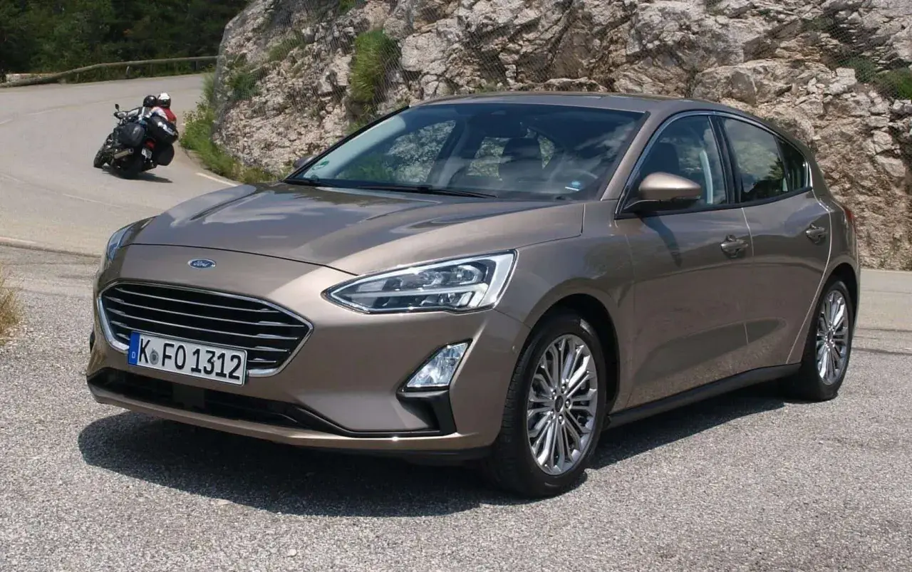 Brązowy Ford Focus 2017 na krętej drodze, z motocyklistą w tle.