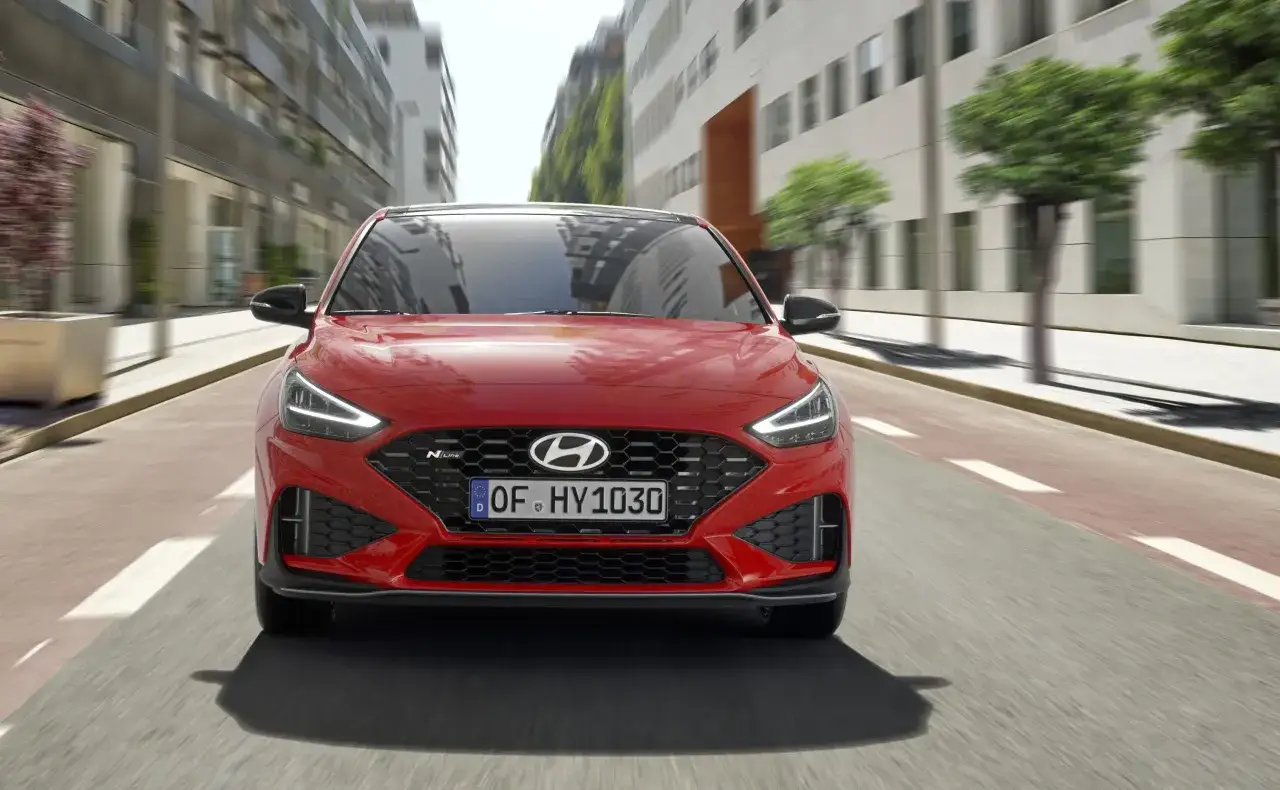 Czerwony Hyundai i30 2023 N Line pędzi ulicą miasta, z budynkami i drzewami w tle.