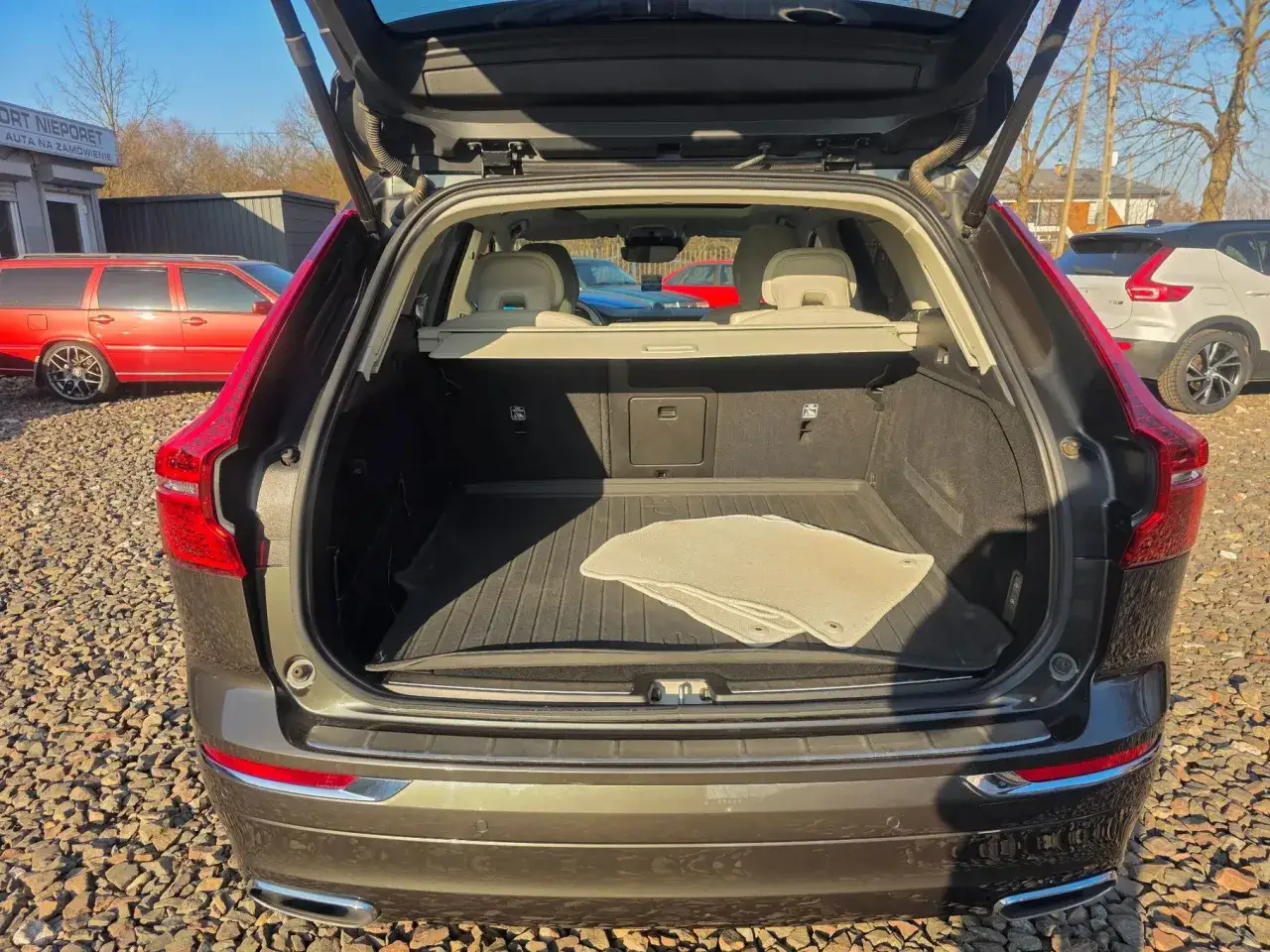 Bagażnik Volvo XC60 T5 otwarty, z widocznymi dywanikami i siatką. W tle inne samochody.