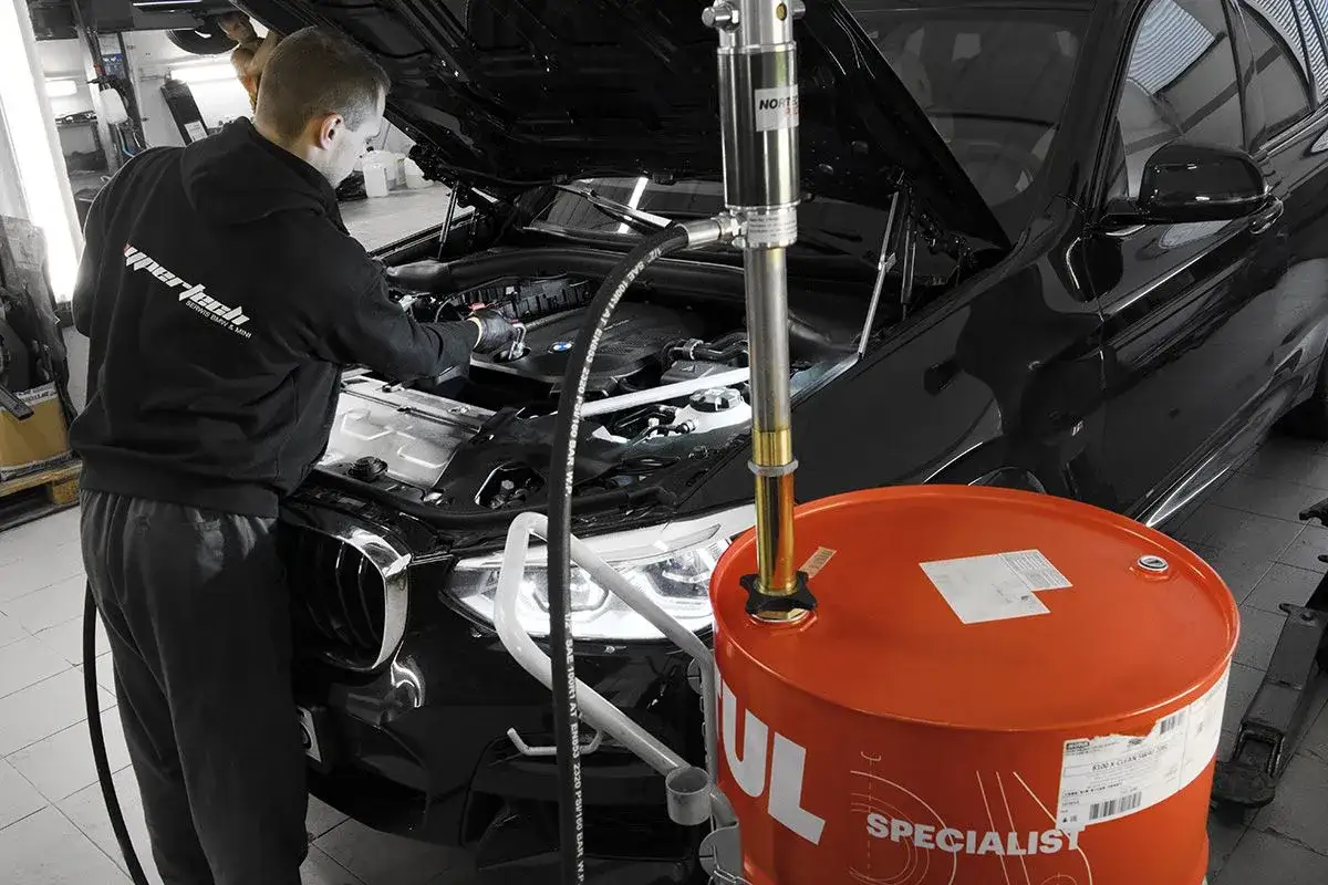 Mechanik wymienia olej w czarnym BMW. Obok stoi duży, pomarańczowy bęben z olejem, przypominając o tym, co ile wymiana oleju jest zalecana.