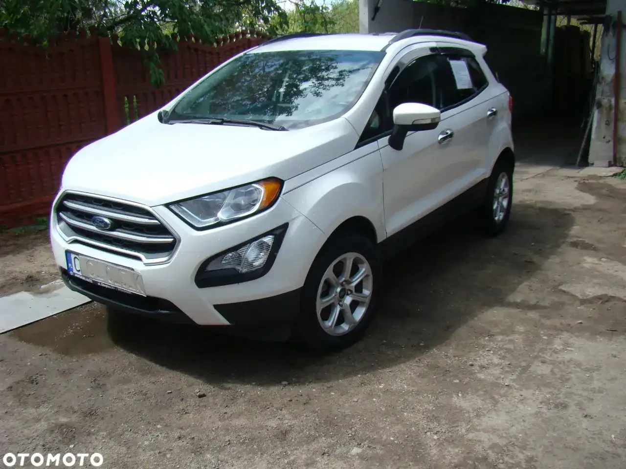 Biały Ford Ecosport zaparkowany na podwórku, obok czerwonego płotu i zadaszenia.
