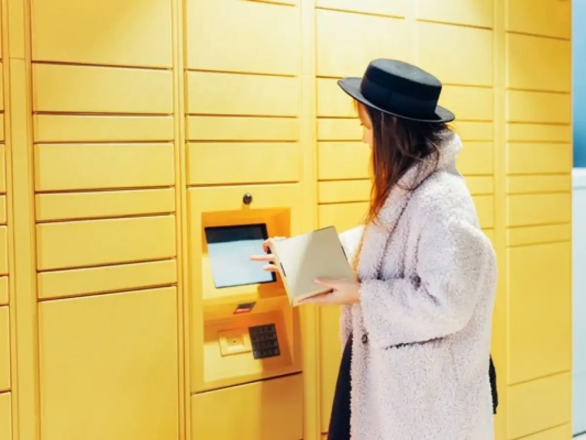 Kobieta w kapeluszu odbiera paczkę magazynowaną InPost z żółtej skrytki.