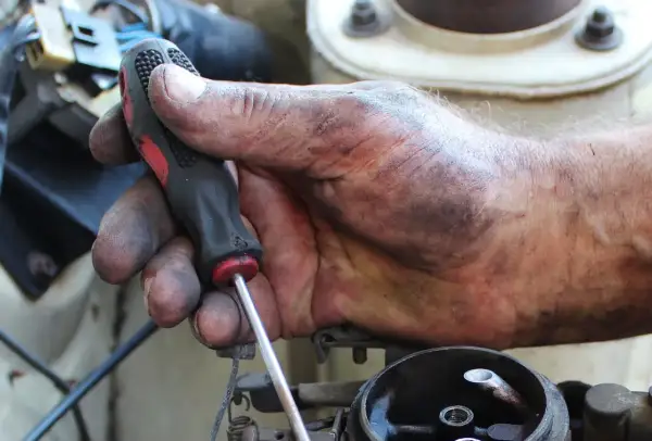 Brudna ręka mechanika trzyma śrubokręt, naprawiając linkę zmiany biegów. Sprawdź wymiana linki zmiany biegów koszt.