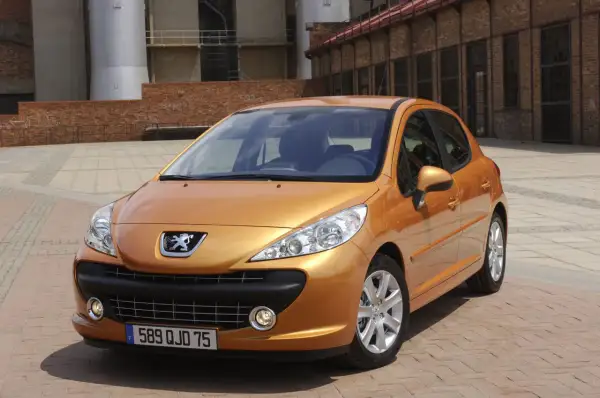 Pomarańczowy Peugeot 207 SW 1.4 benzyna zaparkowany na brukowanym placu przed ceglanym budynkiem.