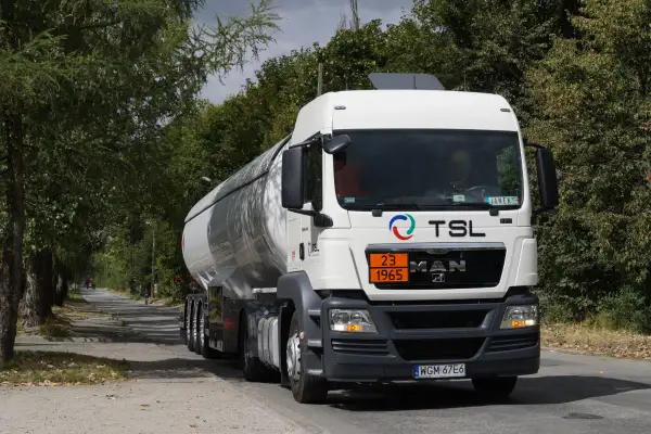 Biały cysterna z logo TSL jedzie drogą wśród drzew. To ważny element branży TSL, transportującej towary.