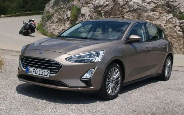 Brązowy Ford Focus 2017 na krętej drodze, z motocyklistą w tle.
